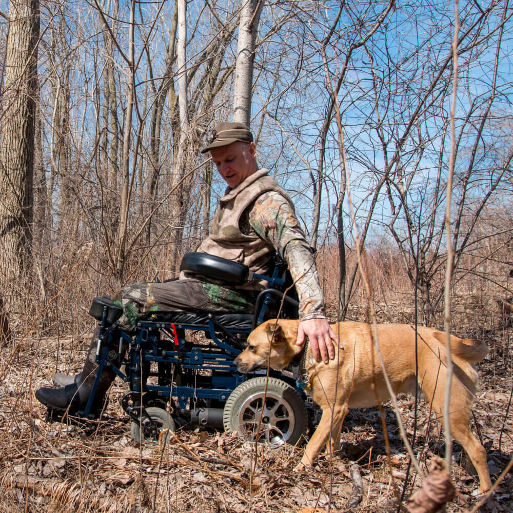 man with mixed power wheelchair in the woods with a dog