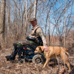 man with mixed power wheelchair in the woods with a dog (thumbnail)